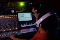 Over the shoulder view of a young mixed race male sound engineer sitting and working at a mixing desk in a recording studio using a computer and wearing headphones and giving a thumbs up sign during a recording session — Stock Photo