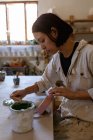 Vue de côté gros plan d'une jeune potière caucasienne assise à une table de travail peignant une glaçure colorée sur une fiole d'argile dans un atelier de poterie — Photo de stock