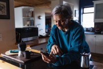 Вид збоку крупним планом старший кавказька жінка на кухні за допомогою смартфона з кухонних шаф у фоновому режимі — стокове фото