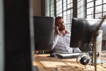 Vue de face gros plan d'un jeune homme caucasien assis à un bureau parlant sur un smartphone dans un bureau créatif, vu entre les écrans d'ordinateur — Photo de stock