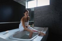 Вид сбоку на молодую брюнетку-кавказку, сидящую на боку ванны и бреющую ноги — стоковое фото
