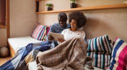Vista frontale di una giovane donna di razza mista e di un giovane afroamericano che guarda un tablet e sorride, seduti insieme su un divano a casa
. — Foto stock