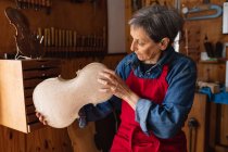 Зовнішній вигляд старших кавказьких жіночих luthier тримає тіло скрипки в її майстерні, з інструментами висить на стіні у фоновому режимі — стокове фото