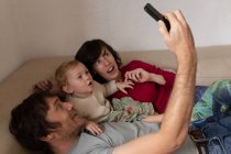 Vista lateral de perto de um jovem pai e mãe caucasianos deitados em um sofá com seu bebê e tirando uma selfie — Fotografia de Stock