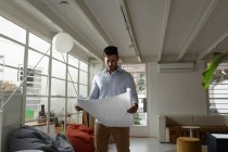 Front view of a young mixed race man standing and checking architectural drawings at a creative office — Stock Photo