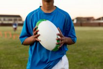 Vue de face du milieu d'une joueuse de rugby debout sur un terrain de sport tenant une balle de rugby lors d'une séance d'entraînement — Photo de stock