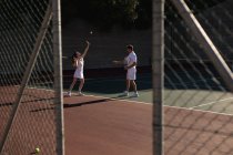 Vue latérale d'une jeune femme caucasienne et d'un homme jouant au tennis par une journée ensoleillée, d'une femme se préparant à servir et d'un homme gesticulant, vus à travers une clôture — Photo de stock