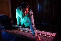 Front view close up of a young Caucasian male sound engineer wearing headphones reaching across a mixing desk in a recording studio to adjust a channel setting — Stock Photo