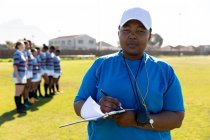 Front view close up of a middle aged mixed race female rugby coach standing on a rugby pitch writing on a clipboard and looking to camera, with her team standing in a row together in the background — Stock Photo