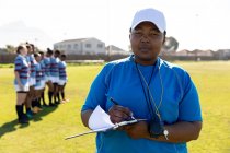 Vista frontale da vicino di un allenatore di rugby femminile di mezza età in piedi su un campo da rugby che scrive sugli appunti e guarda alla telecamera, con la sua squadra in fila sullo sfondo — Foto stock