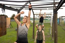 Front view of a young Caucasian woman hanging from monkey bars at an outdoor gym during a bootcamp training session with two other participants in the background — Stock Photo