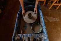 Vista elevada de las manos de una alfarera sosteniendo una rueda de alfarero con una olla en ella en un estudio de cerámica - foto de stock