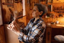 Seitenansicht einer älteren kaukasischen Geigenbauerin mit der Rolle einer Geige in ihrer Werkstatt, im Hintergrund hängen Werkzeuge an der Wand — Stockfoto