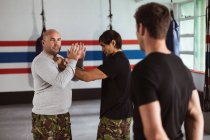 Side view close up of a middle aged Caucasian male instructor giving self defence training in a boxing gym demonstrating a hold on a young mixed race man, while another young man looks on — Stock Photo