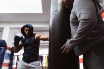 Nahaufnahme eines jungen Boxer mit gemischter Rasse, der in einer Boxhalle einen Boxsack schlägt, der von einem kaukasischen Trainer mittleren Alters gehalten wird — Stockfoto