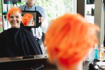 Vista trasera de cerca de una peluquería caucásica de mediana edad y una joven mujer caucásica que tiene su cabello de color rojo brillante y se muestra en un espejo de mano en un salón de belleza - foto de stock