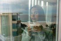 Вид спереду крупним планом молодий Кавказький жінка брюнетка одягнений в сірий светр, стоячи і дивлячись з вікна, тримаючи чашку кави, побачити через вікно з відображенням будівель за межами — стокове фото