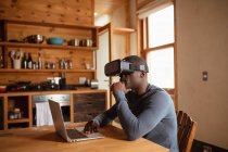 Vista lateral de cerca de un joven afroamericano con auriculares VR usando un portátil sentado en la mesa de la cocina en casa - foto de stock