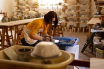 Vista lateral de una joven alfarera caucásica sentada y trabajando con arcilla sobre una rueda de alfarero en un estudio de cerámica, con equipo en primer plano - foto de stock