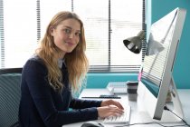 Seitenansicht einer jungen kaukasischen Frau, die an einem Schreibtisch am Fenster sitzt und einen Computer benutzt, sich im modernen Büro eines kreativen Unternehmens der Kamera zuwendet und lächelt — Stockfoto
