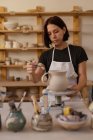 Vista frontal close-up de uma jovem cerâmica caucasiana em uma mesa de trabalho vidros um jarro em uma roda de banda em um estúdio de cerâmica, com vasos e ferramentas em primeiro plano — Fotografia de Stock