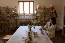 Вид сбоку на молодую кавказскую гончарку, сидящую за рабочим столом и рисующую цветную глазурь на глиняной фляжке в керамической мастерской — стоковое фото