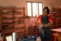 Vista frontale di una giovane ceramista caucasica che si pulisce le mani sul grembiule sporco che indossa, in piedi accanto a una ruota di vasai in uno studio di ceramica — Foto stock