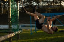 Вид сбоку на молодого кавказца без рубашки, висящего на горизонтальной веревке на скалолазании в открытом спортзале во время тренировочного лагеря — стоковое фото