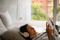 Seitenansicht einer jungen kaukasischen brünetten Frau, die mit Kopfhörern auf einem Sofa liegt und einen Tablet-Computer anschaut — Stockfoto