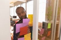 Vista frontal close-up de um jovem afro-americano escrevendo notas em uma parede de vidro durante uma sessão de brainstorm em equipe em um escritório criativo, visto através da parede de vidro — Fotografia de Stock