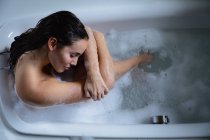 Overhead close up de uma jovem mulher morena caucasiana sentada em um banho de espuma, segurando suas pernas desenhadas e descansando a cabeça em seus braços com os olhos fechados — Fotografia de Stock