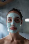 Porträt einer jungen kaukasischen Brünetten mit Gesichtsmaske, die in einem modernen Badezimmer direkt in die Kamera blickt — Stockfoto