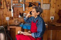 Vista lateral de uma mulher caucasiana sênior luthier falando ao telefone em sua oficina com ferramentas penduradas na parede no fundo — Fotografia de Stock