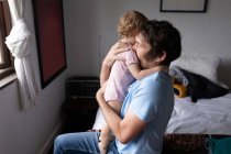 Vista laterale di un giovane padre caucasico che tiene in braccio il suo bambino, seduto su un letto — Foto stock