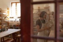 Vista lateral de uma jovem cerâmica caucasiana que inspeciona um prato em um estúdio de cerâmica, vista através de uma porta de vidro — Fotografia de Stock