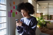 Side view close up of a young mixed race woman sitting on an armchair and using a smartphone at a creative office — Stock Photo