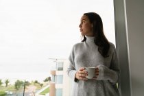 Front view of a young Caucasian brunette woman wearing a grey turtleneck sweater, standing on a balcony holding a cup of coffee with her head turned to the side, looking away — Stock Photo