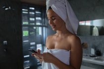 Vista lateral de perto de uma jovem morena caucasiana vestindo uma toalha de banho e com o cabelo embrulhado em uma toalha, usando um smartphone em um banheiro moderno — Fotografia de Stock