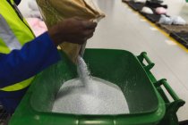 Visão lateral seção média do trabalhador da fábrica masculino derramando um grânulos brancos de um saco em um recipiente de plástico verde para um balde em um armazém em uma fábrica de processamento — Fotografia de Stock