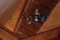 Vista aérea de dos niños caucásicos preadolescentes sentados en una escalera en casa, usando una tableta y un teléfono inteligente - foto de stock