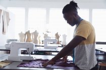 Vue latérale d'un jeune étudiant afro-américain en mode masculine coupant du tissu tout en travaillant sur un design dans un studio au collège de mode — Photo de stock