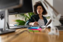 Vue de face gros plan d'une jeune femme métisse assise à un bureau regardant un écran d'ordinateur et utilisant un stylet dans un bureau créatif — Photo de stock