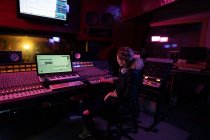 Vista lateral de una joven ingeniera de sonido caucásica sentada y trabajando en un escritorio de mezclas en un estudio de grabación rodeado de equipo - foto de stock