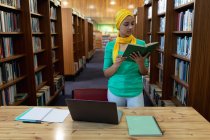 Передній погляд на молоду азіатську студентку, одягнену в хіджаб, яка читає книжку, користується лептопом і навчається в бібліотеці. — стокове фото