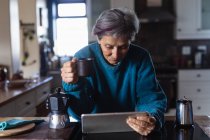 Вид на старших кавказька жінка в кухні пити каву і за допомогою планшетного комп'ютера з кухонних шаф у фоновому режимі — стокове фото