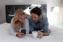 Вид на щасливу молоду Кавказьку пару разом відпочиваючи на відпочинку в готельному номері лежачи на ліжку за допомогою смартфона — стокове фото