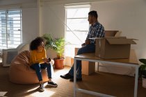 Side view close up of a young mixed race man sitting on a desk and a young mixed race woman sitting on a bean bag having a discussion with cardboard boxes at a creative office — Stock Photo