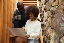 Front view close up of a young mixed race woman and a young African American man looking at a laptop computer and talking sitting on the stairs at home. — Stock Photo