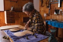 Seitenansicht einer älteren kaukasischen Geigenbauerin, die in ihrer Werkstatt am Korpus einer Geige arbeitet, im Hintergrund hängen Werkzeuge an der Wand — Stockfoto
