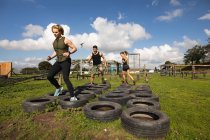 Front view of two young Caucasian women and a young Caucasian man stepping through tyres at an outdoor gym during a bootcamp training session — Stock Photo
